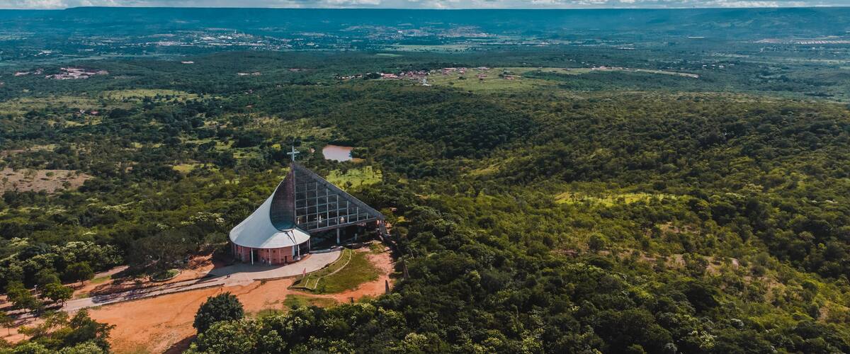 A church with a distinctive roof and a cross atop, surrounded by lush greenery and a vast landscape.