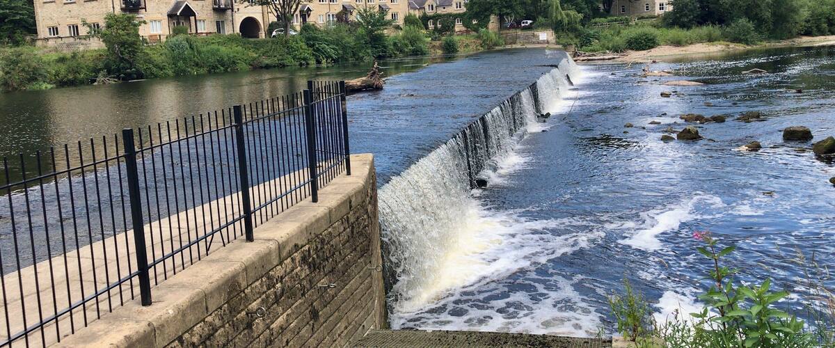 A river with a weir, a stone bridge, and residential buildings in the background.