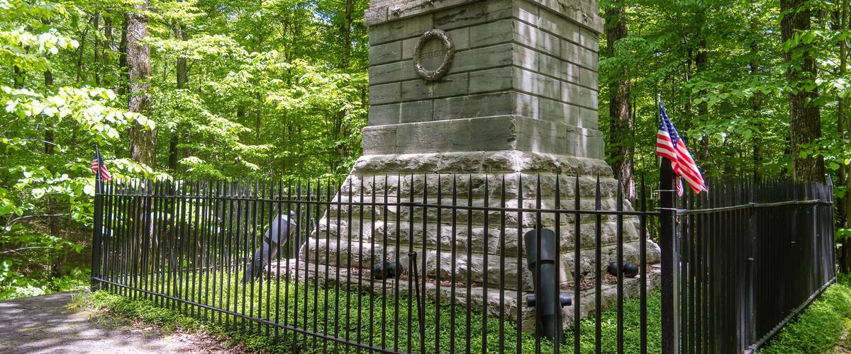 A stone monument with an American flag, surrounded by a metal fence and lush green trees.