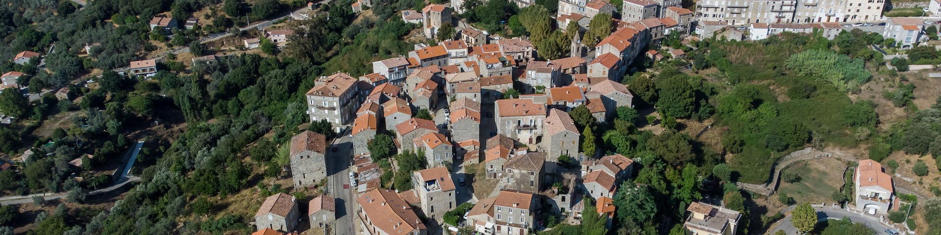 An aerial view of a densely built village with stone buildings and terraced houses, surrounded by lush greenery and hills.