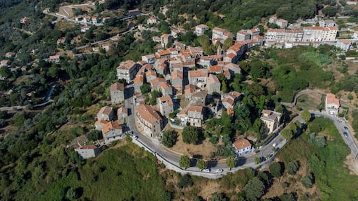An aerial view of a densely built village with stone buildings and terraced houses, surrounded by lush greenery and hills.