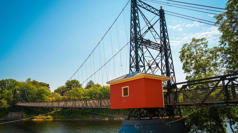 A suspension bridge with a red structure on the right side, a river below, and trees in the background.