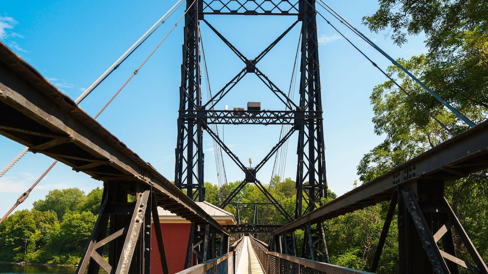 A steel bridge with a truss design, surrounded by lush green trees and a body of water.