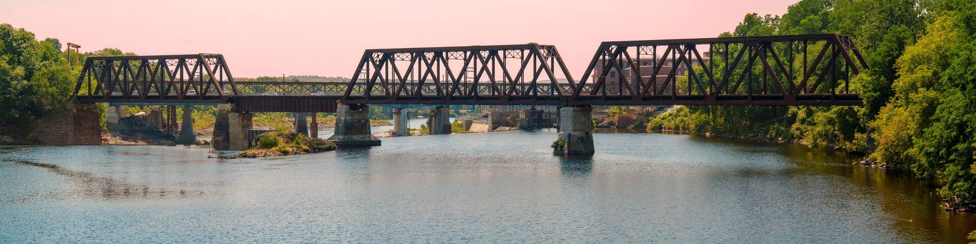 Rustic railway tracks and the historic Waterville–Winslow Railroad Bridge span the Kennebec River in Waterville, Maine, USA—a steel truss structure built around natural rock formations