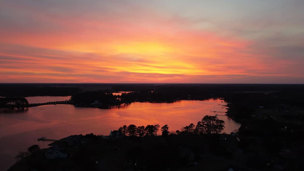 A sunset over a calm lake with a bridge and silhouetted trees.