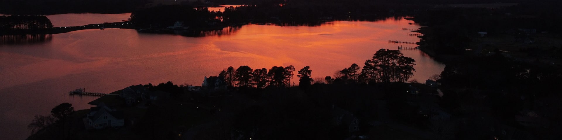 A sunset over a calm lake with a bridge and silhouetted trees.