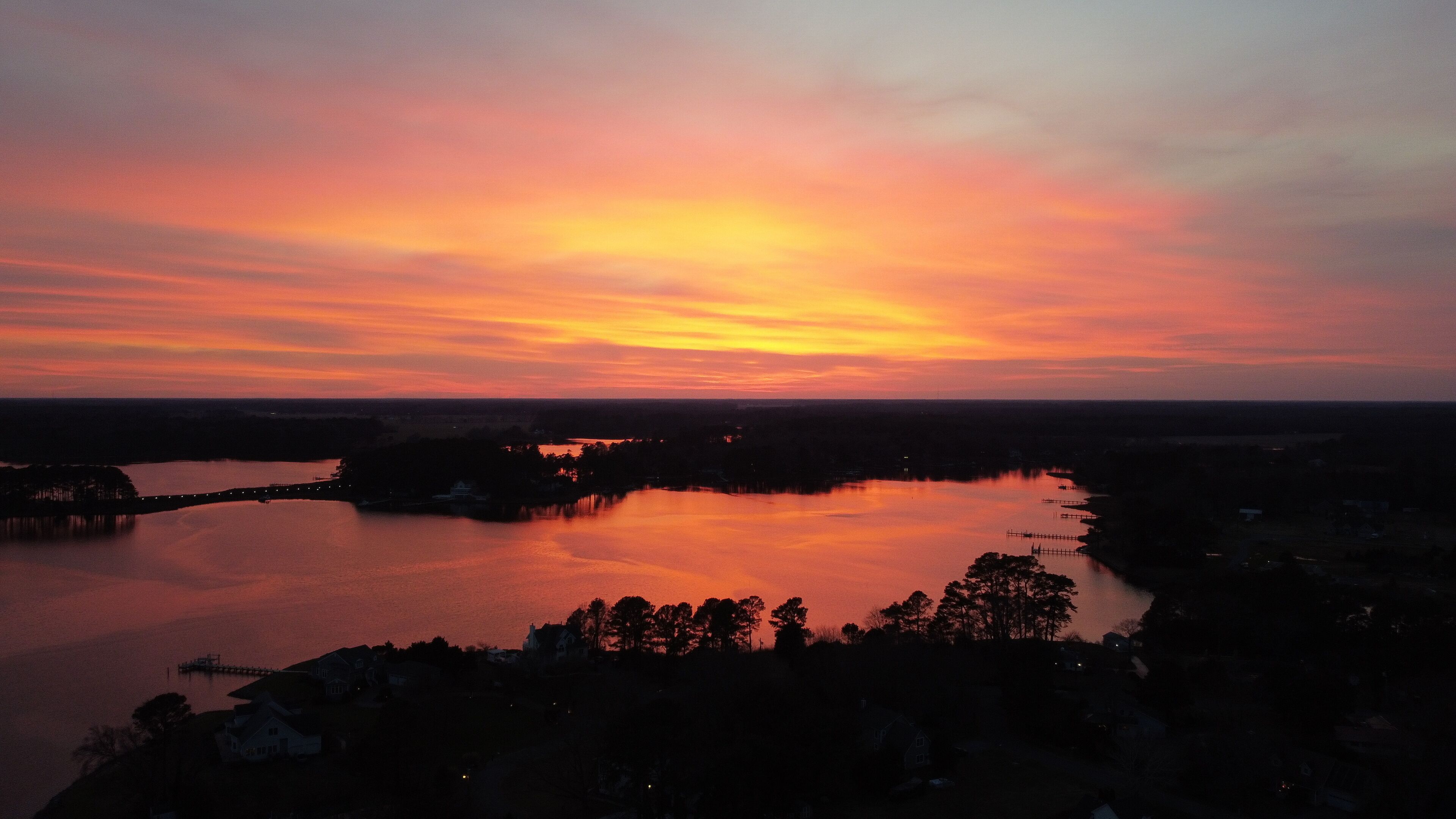 A sunset over a calm lake with a bridge and silhouetted trees.