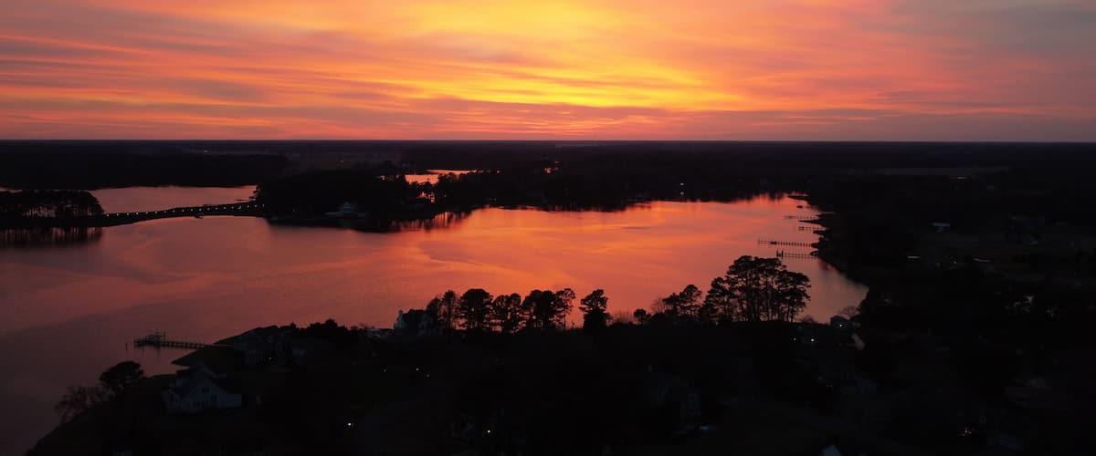 A sunset over a calm lake with a bridge and silhouetted trees.
