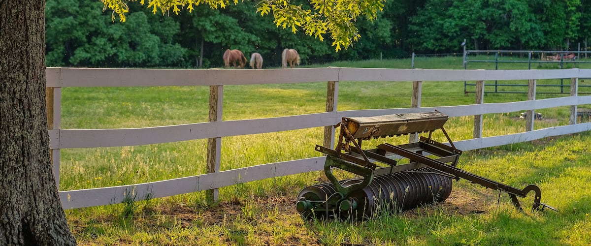 A white wooden fence, a tree, and a tractor in a grassy field with horses in the background.