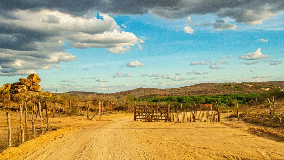 A dirt road with a wooden gate, surrounded by sparse vegetation and hills in the background.