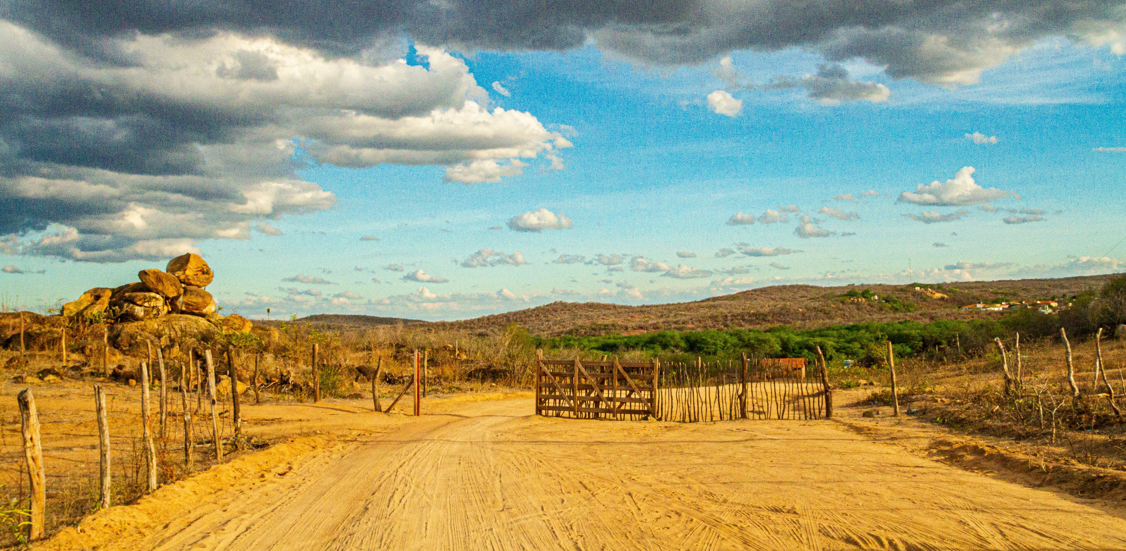 A dirt road with a wooden gate, surrounded by sparse vegetation and hills in the background.