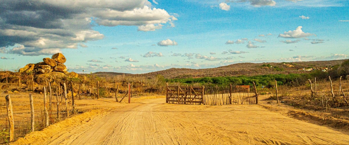 A dirt road with a wooden gate, surrounded by sparse vegetation and hills in the background.
