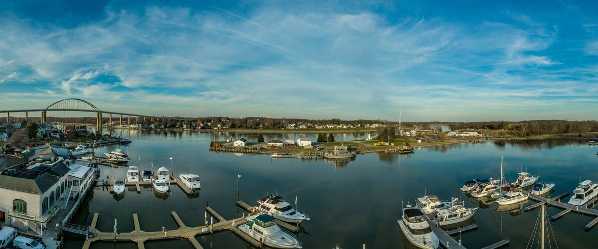 A marina with boats docked, a bridge in the background, and a calm river.