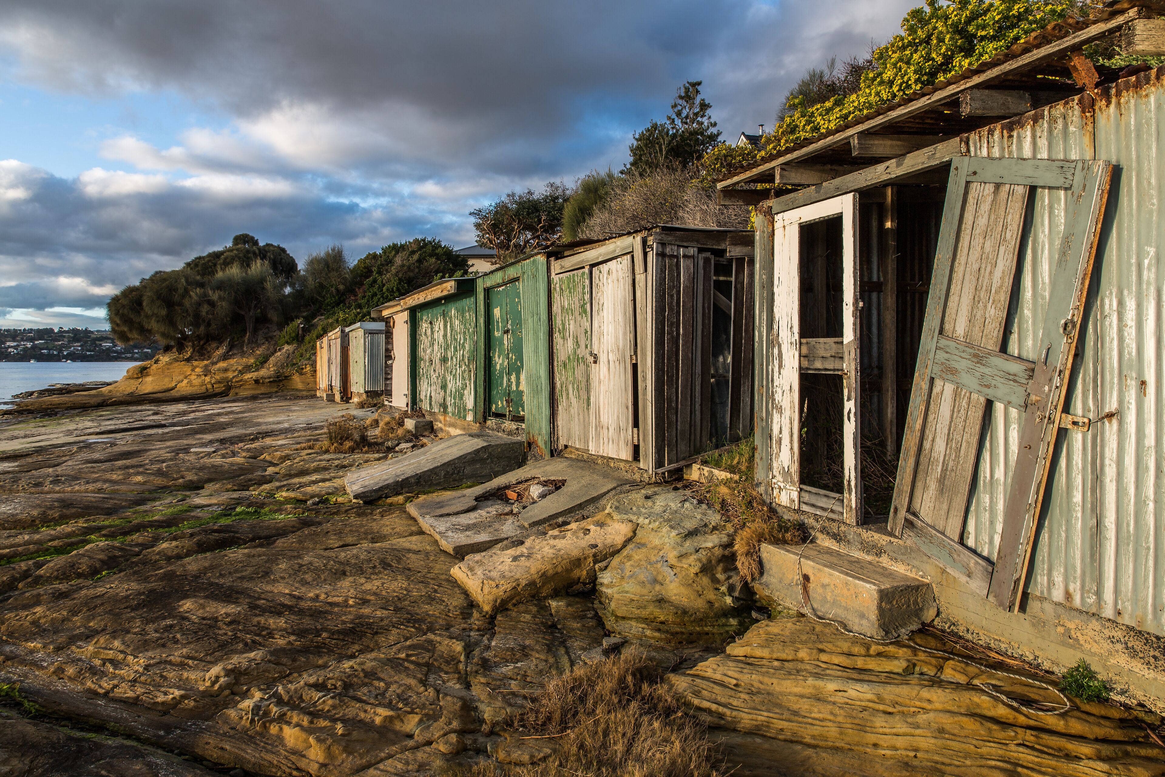 A row of weathered wooden buildings with green doors, situated on a rocky shoreline with a view of the sea and distant hills.
