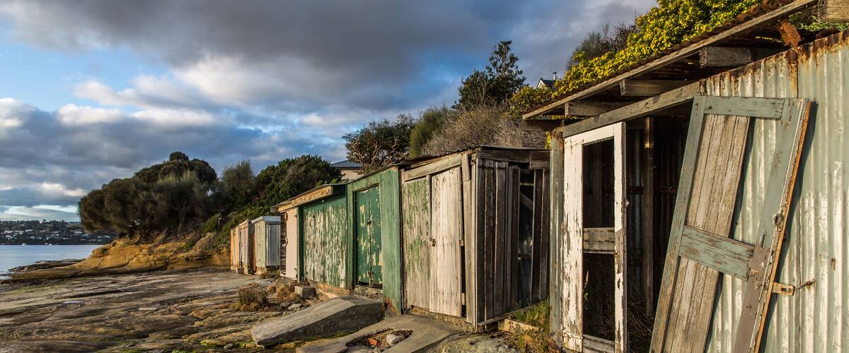 A row of weathered wooden buildings with green doors, situated on a rocky shoreline with a view of the sea and distant hills.
