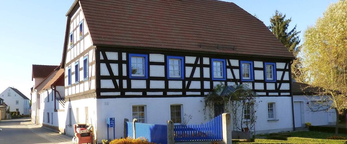 A traditional half-timbered house with a red-tiled roof and blue window frames, surrounded by a cobblestone street and orange flowers.
