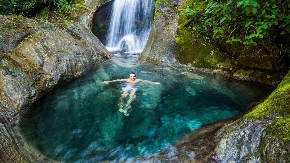 A person swimming in a clear, rocky pool with a waterfall in the background.