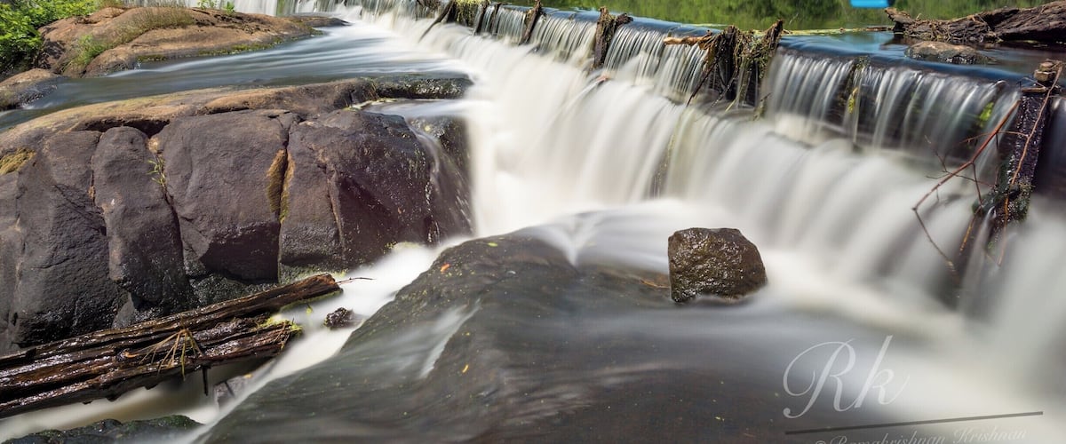 A waterfall cascading over rocks with a forested background and a clear sky.