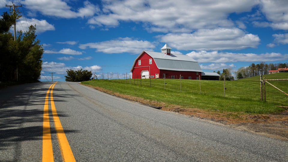 A red barn with a white door and a grey roof, surrounded by green fields and a clear blue sky with clouds.