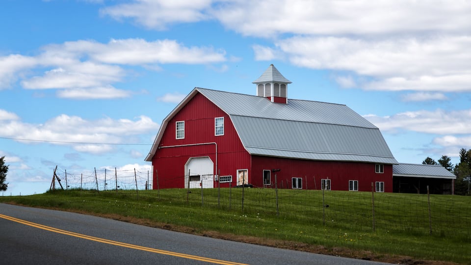 A red barn with a white door and a metal roof, surrounded by green fields and a road.