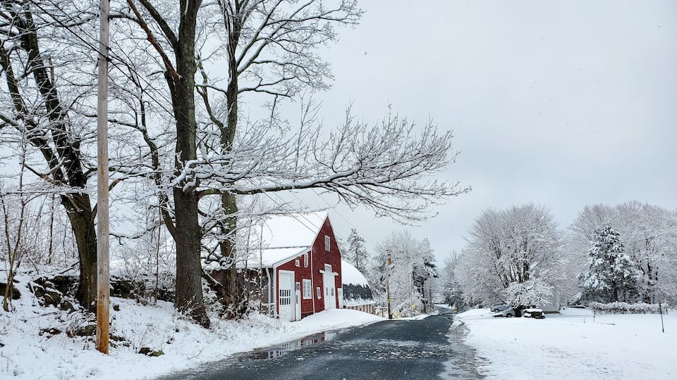 A snow-covered rural road with a red barn and leafless trees.
