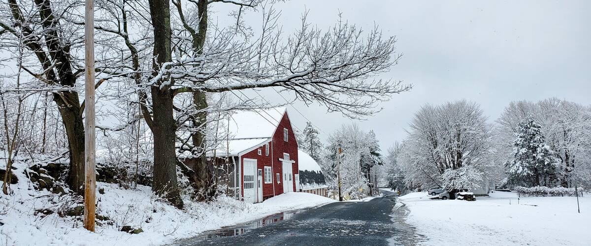 A snow-covered rural road with a red barn and leafless trees.