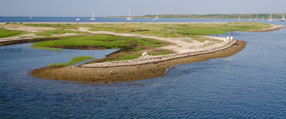 A coastal landscape with a sandy beach, grassy areas, and a small island with a lighthouse.