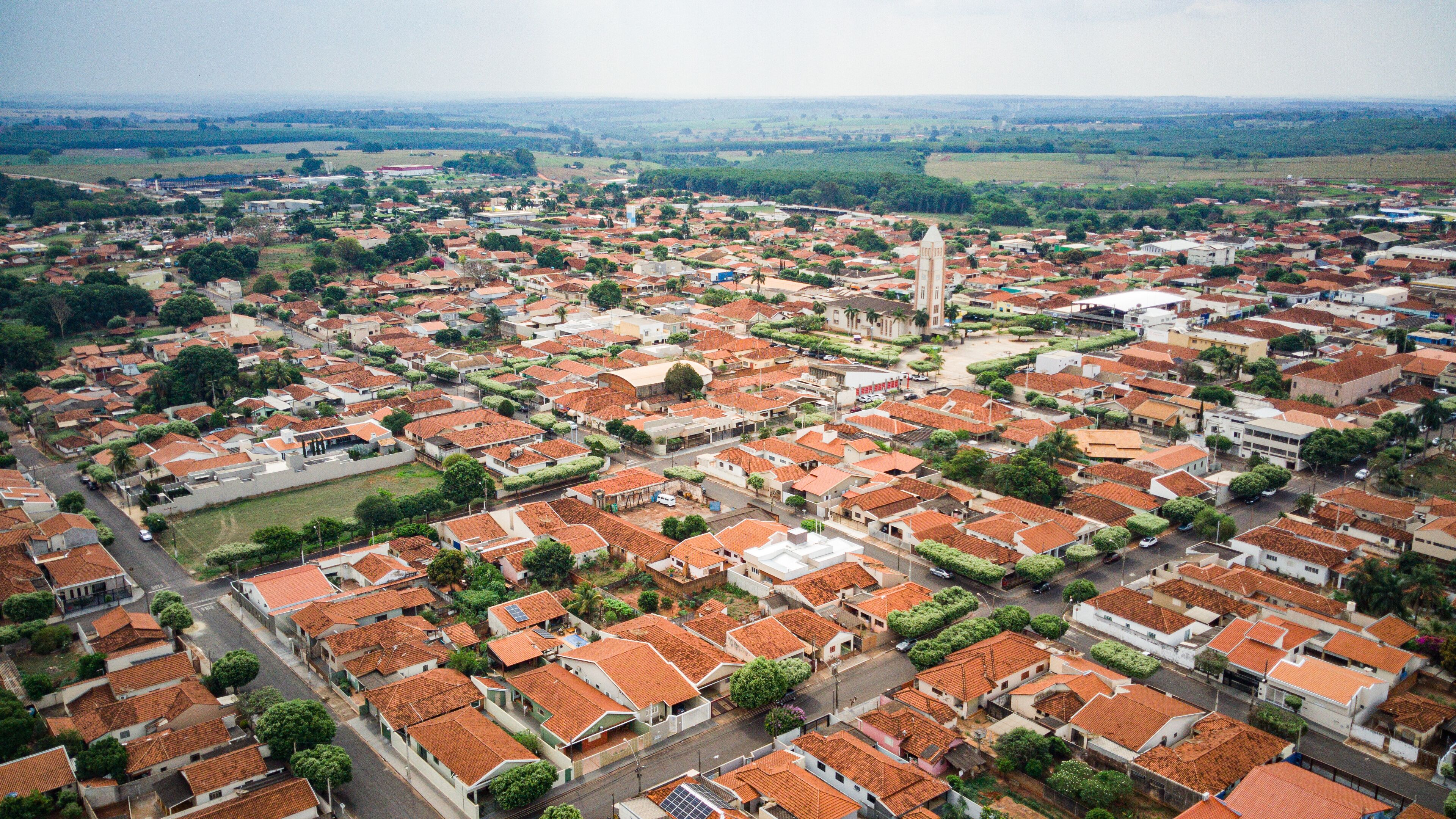 An aerial view of a town with red-tiled roofs, a prominent church tower, and surrounding green fields.