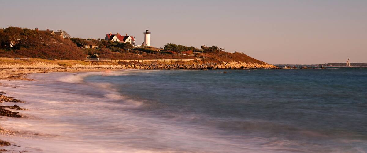 A coastal landscape with a lighthouse, beach, and distant bridge.