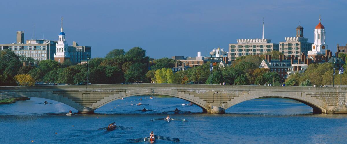A stone bridge over a river with rowing activity, surrounded by trees and buildings with distinctive architecture.