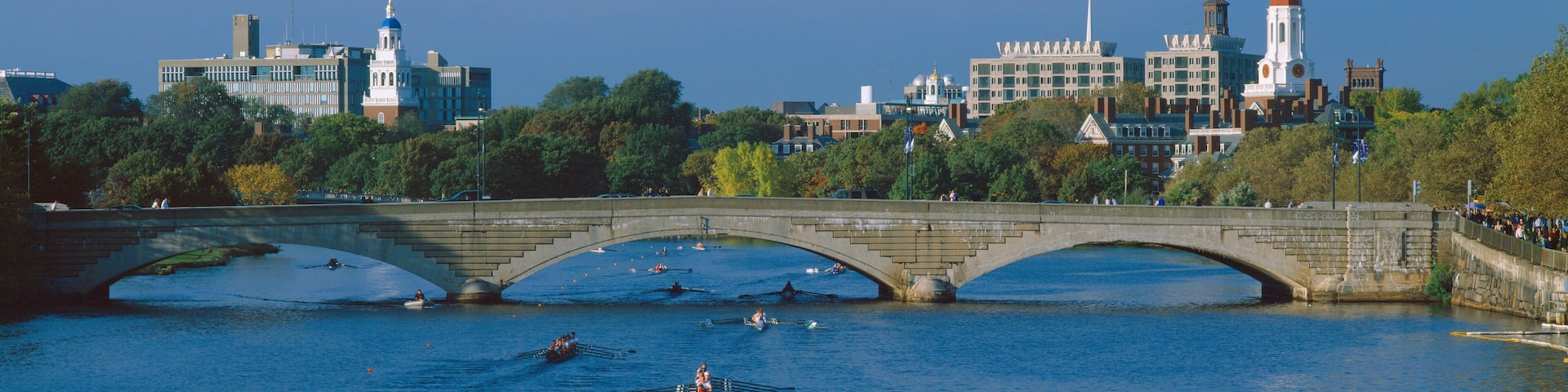 A stone bridge over a river with rowing activity, surrounded by trees and buildings with distinctive architecture.
