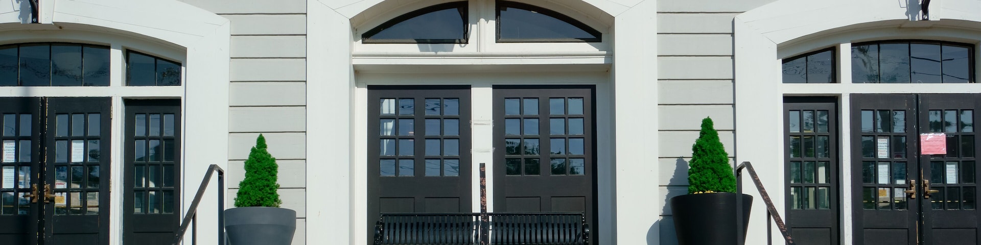Un ayuntamiento con un cartel sobre la entrada, una escalinata que lleva a las puertas y dos árboles en macetas a cada lado.