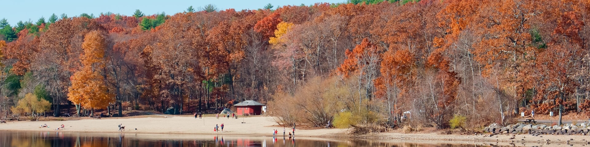 A calm lake reflecting autumn trees, a sandy beach, and a small red building.