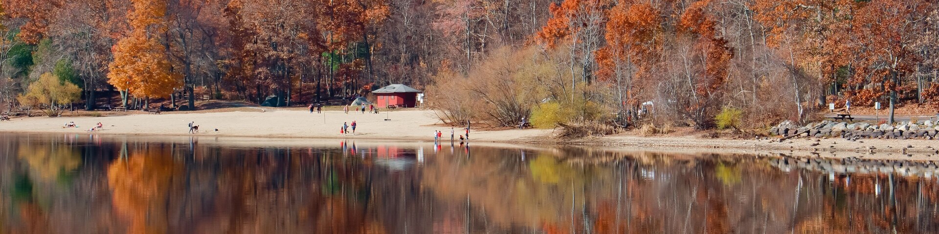 Un lago tranquilo que refleja árboles otoñales, una playa arenosa y un pequeño edificio rojo.