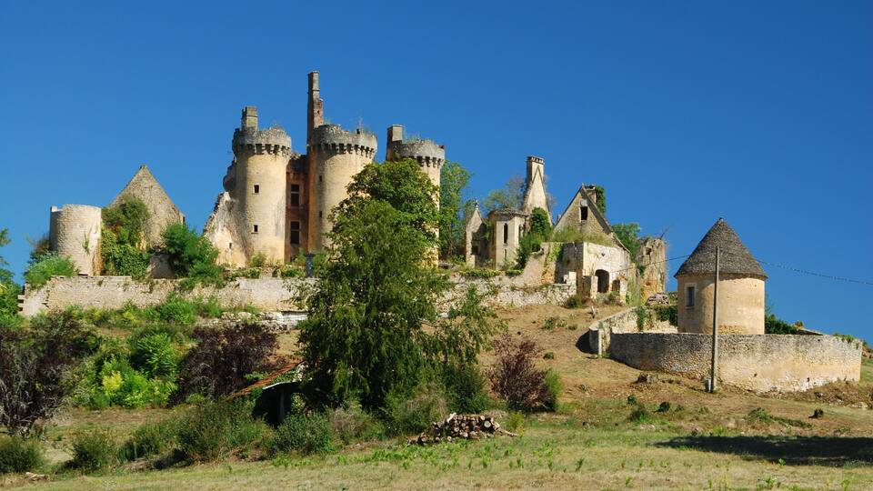 A historic castle with multiple towers and turrets, surrounded by greenery and a clear blue sky.