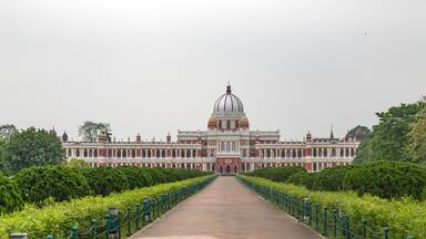 A grand building with a large dome, a long central pathway, and green hedges on both sides.