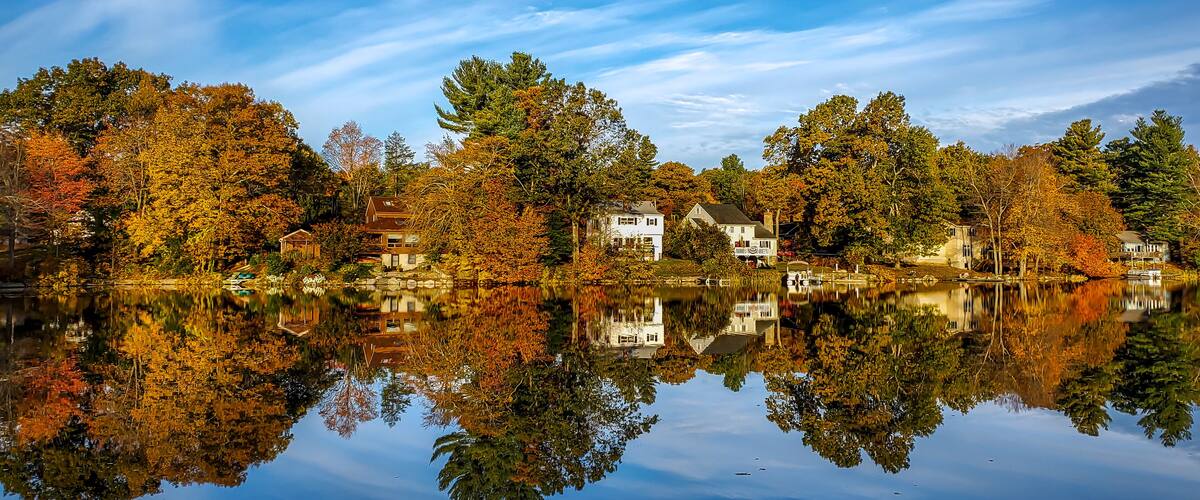 A serene lake with autumn-colored trees reflecting on the water, surrounded by houses.