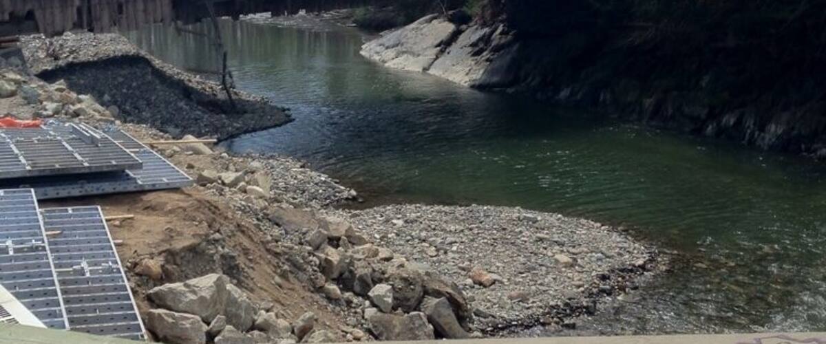 A wooden bridge over a river, solar panels, and a rocky shoreline.