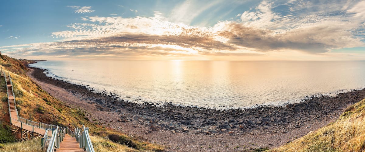 A coastal landscape with a wooden staircase leading down to a rocky beach, a calm sea, and a sunset sky.