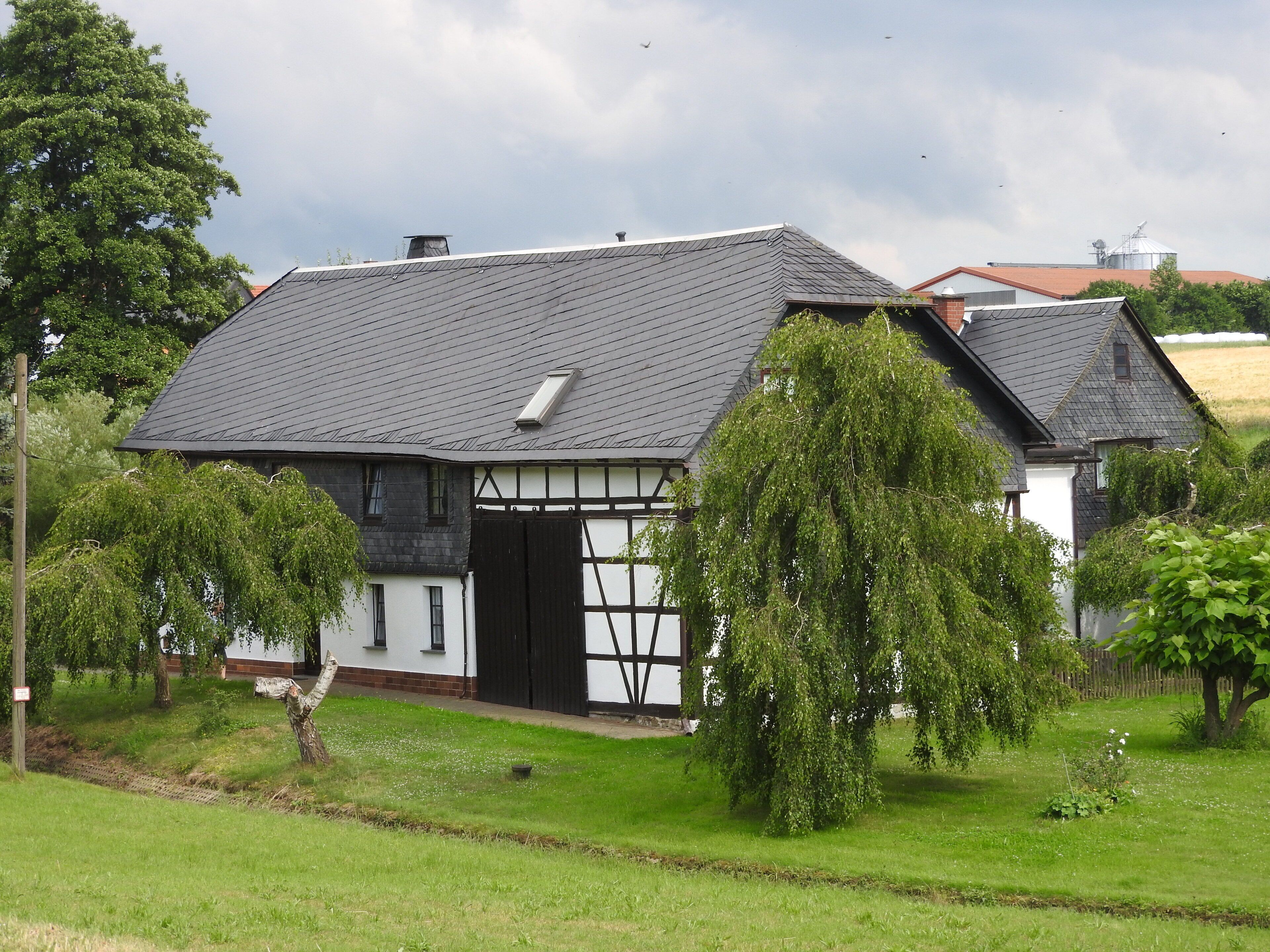 A traditional half-timbered house with a sloped roof, surrounded by lush greenery and trees.