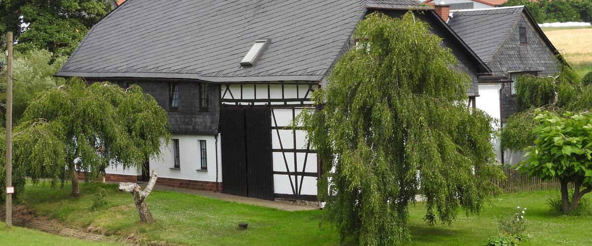A traditional half-timbered house with a sloped roof, surrounded by lush greenery and trees.