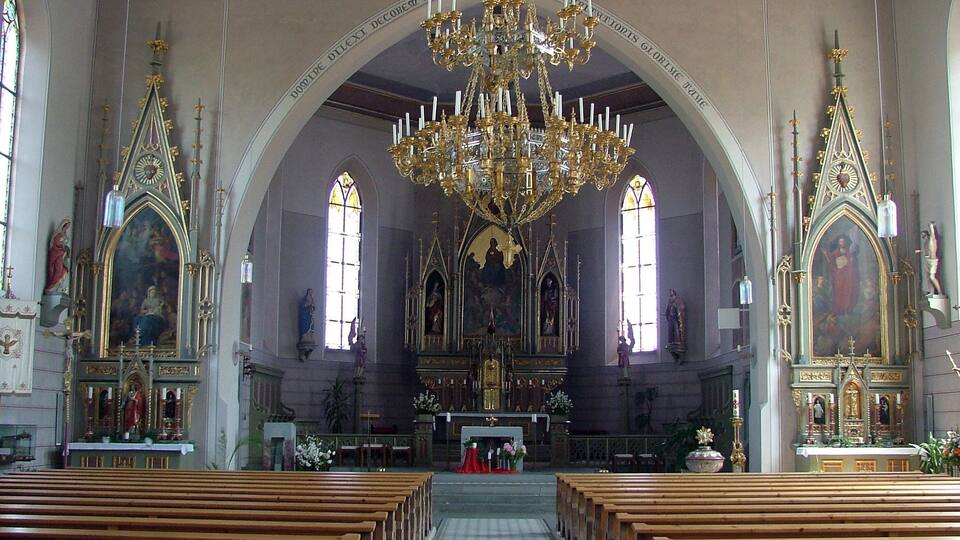 An ornate church interior with a large chandelier, arched windows, and religious paintings.