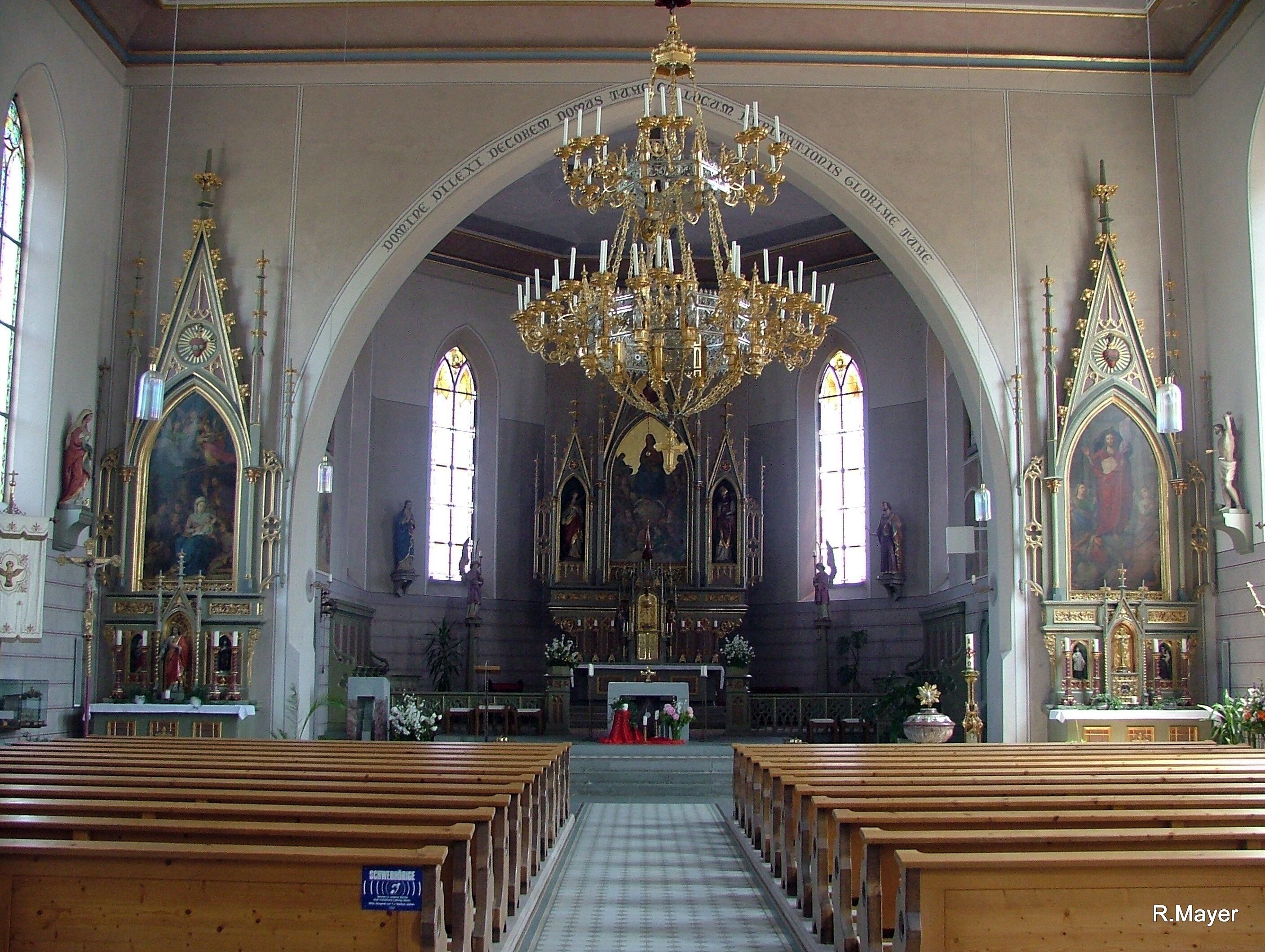 An ornate church interior with a large chandelier, arched windows, and religious paintings.