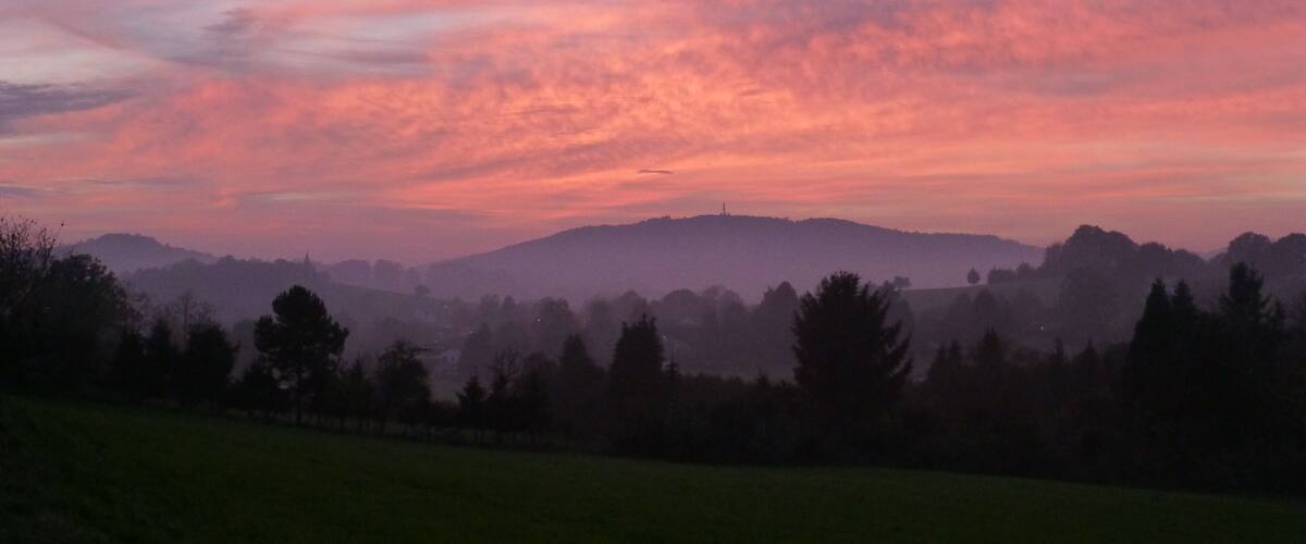 A sunset over a hilly landscape with a field in the foreground and trees silhouetted against the sky.