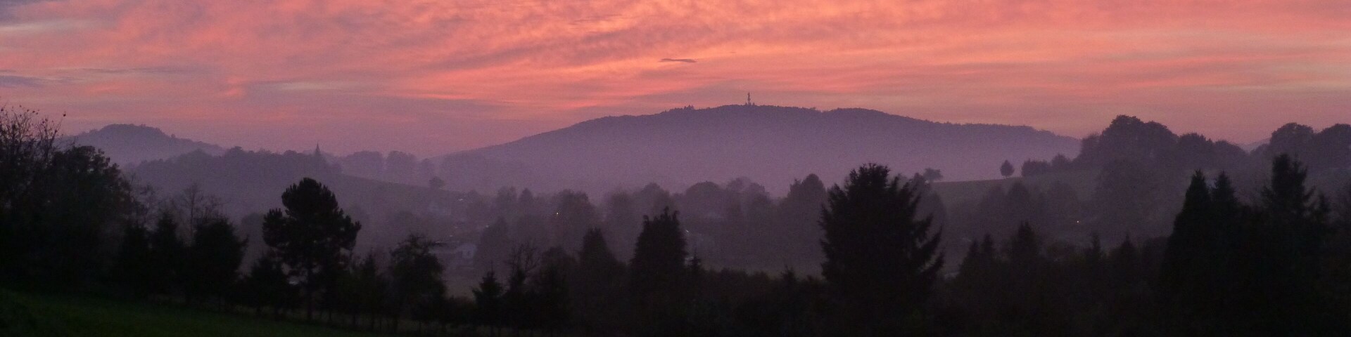 A sunset over a hilly landscape with a field in the foreground and trees silhouetted against the sky.