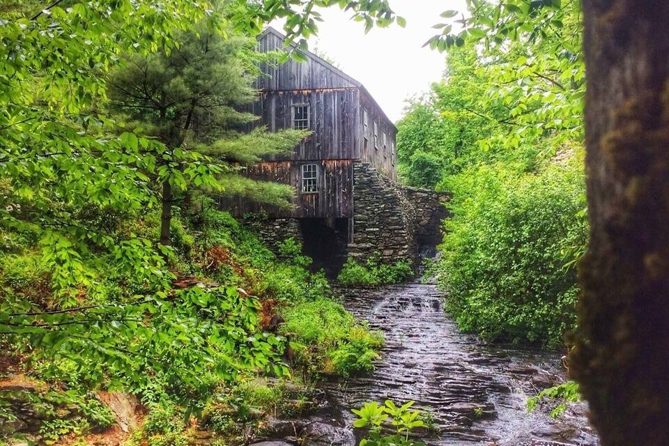 A rustic wooden house with a stone bridge over a flowing stream, surrounded by lush greenery.