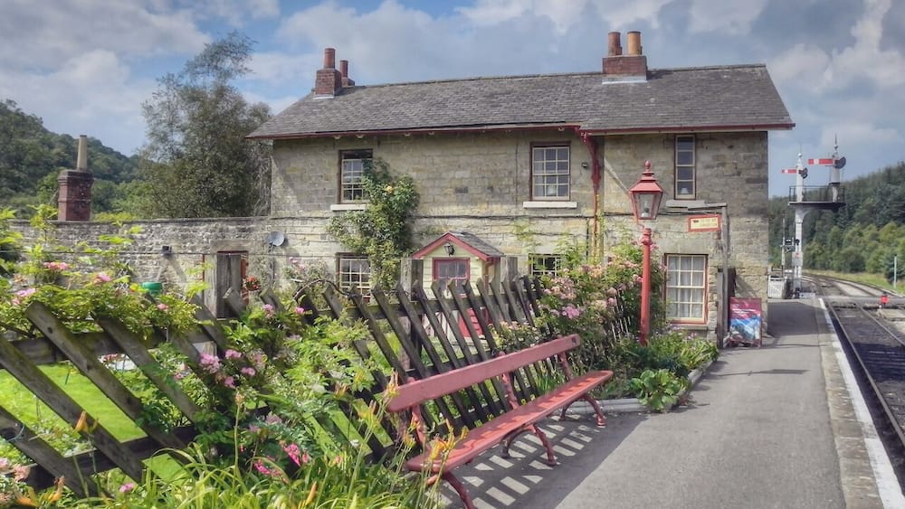 Un bâtiment en pierre avec un banc rouge, entouré d’une végétation luxuriante et situé près d’un passage à niveau.