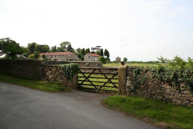 A rural road with a stone wall and a wooden gate leading to a house with a red roof.
