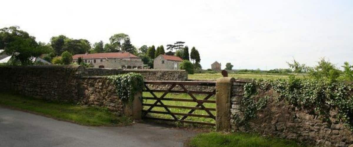 A rural road with a stone wall and a wooden gate leading to a house with a red roof.
