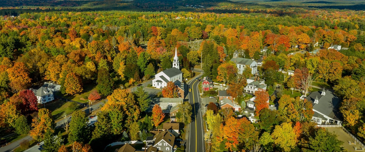 An aerial view of a town surrounded by autumn-colored trees, with a prominent church and residential houses.